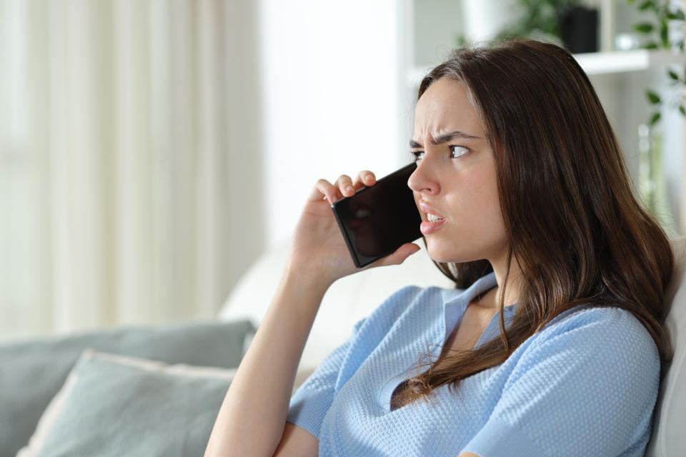 A woman with long hair is sitting on a couch, holding a phone to her ear, looking concerned