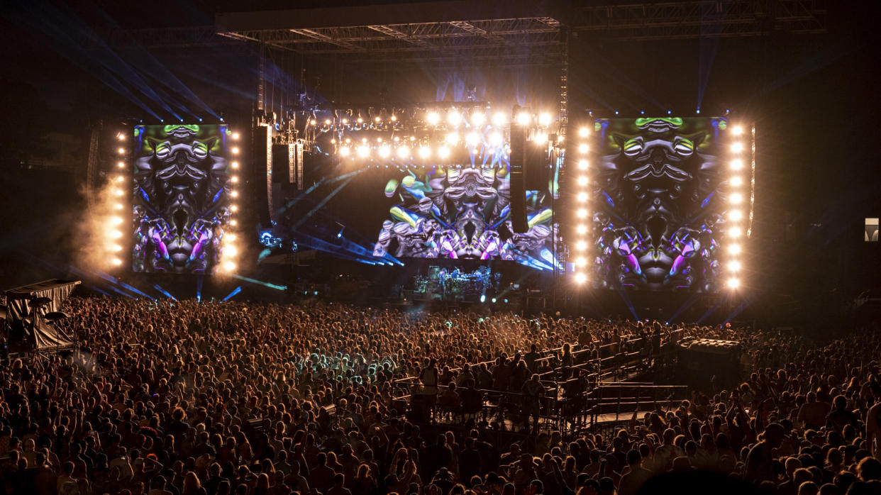  Dead & Co perform at night on a stage with bright lights and colors as onlooking Deadheads dance. 