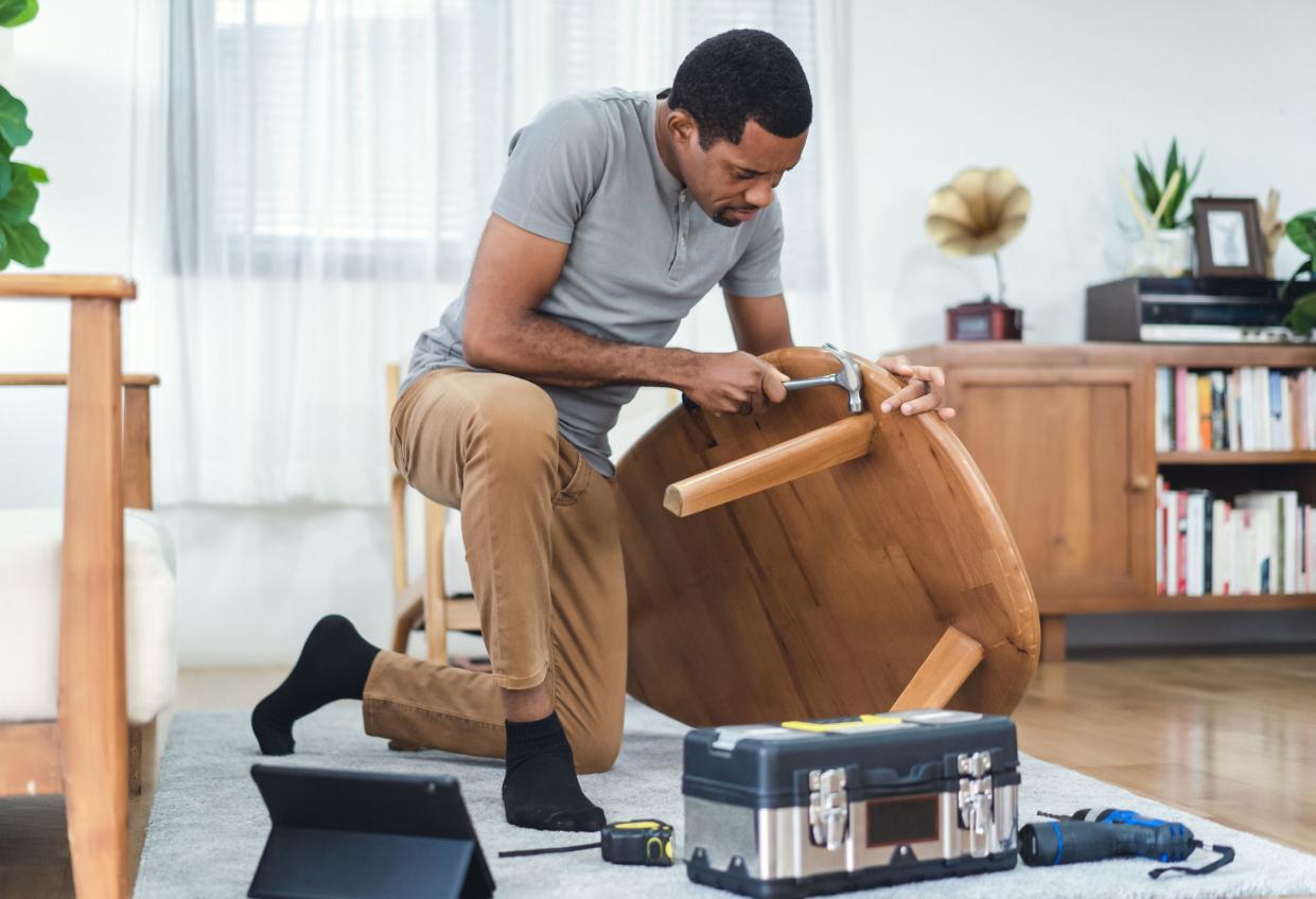 Happy bearded African American man watching online tutorial instructions for assembling furniture about repairing of wooden table. Black man looking on digital tablet screen and hammer assembling.