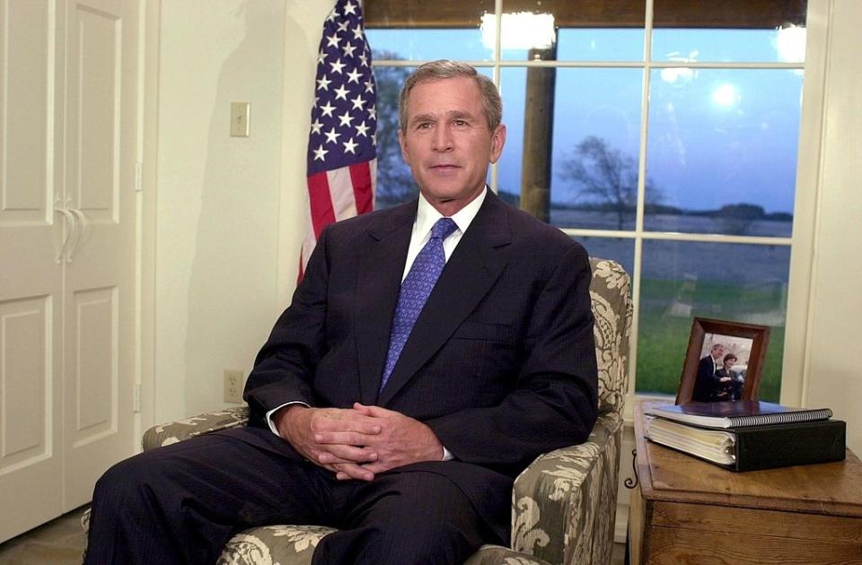 George W. Bush seated in an armchair with hands clasped, in front of an American flag and a window, with a family photo and a binder on a side table