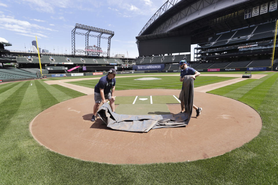 Grounds crew workers Jacob Weiderstrom, left, and Marcus Gignac pull a tarp off home plate as they continue to keep the Seattle Mariners' field in playing shape as the ballpark goes into its seventh week without baseball played because of the coronavirus outbreak Monday, May 11, 2020, in Seattle. A person familiar with the decision tells The Associated Press that Major League Baseball owners have given the go-ahead to making a proposal to the players' union that could lead to the coronavirus-delayed season starting around the Fourth of July weekend in ballparks without fans. (AP Photo/Elaine Thompson)