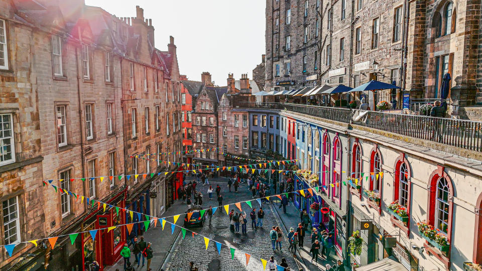 A busy street scene in a historic town with colorful storefronts, hanging flags, and people walking along the cobblestone path