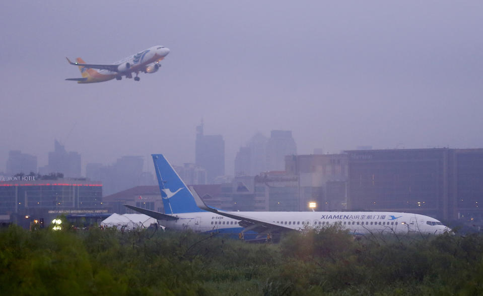 A Cebu Pacific passenger plane takes off, in background, while a Boeing passenger plane from China, a Xiamen Air, lies on the grassy portion of the runway of the Ninoy Aquino International Airport after it skidded off the runway while landing Friday, Aug. 17, 2018 in suburban Pasay city southeast of Manila, Philippines. All the passengers and crew of Xiamen Air flight MF8667 were safe and were taken to an airport terminal, where they were given blankets and food before being taken to a hotel. (AP Photo/Bullit Marquez)