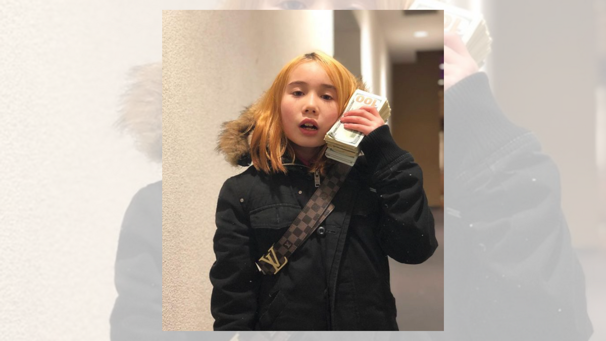 A white girl with blond hair wearing a black jacket and a stack of cash next to her right cheek is standing in a hallway. Image Via @liltay/Instagram