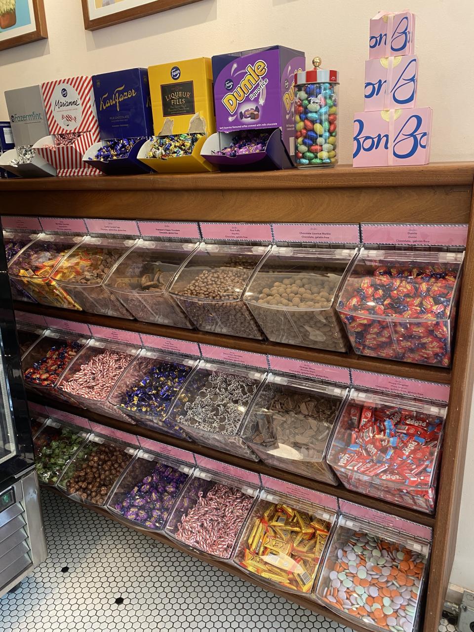 Candy display shelves filled with various candies in clear bins and boxes. Boxes on top include brands like Lemon Bon Bons, Dumle, and others