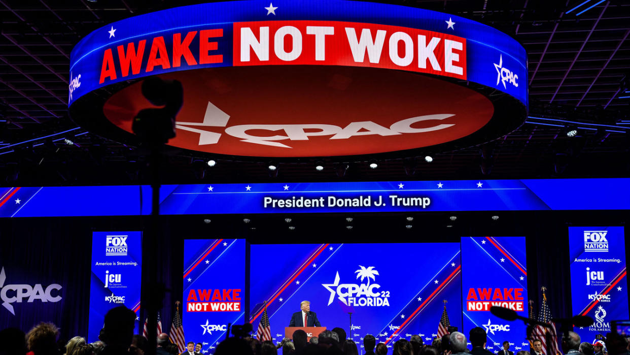Donald Trump at the podium under a huge sign that says in red, white and blue: Awake Not Woke.