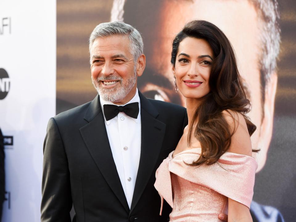 George Clooney wearing a tuxedo and a bowtie with Amal Clooney wearing a pink off-the-shoulder dress