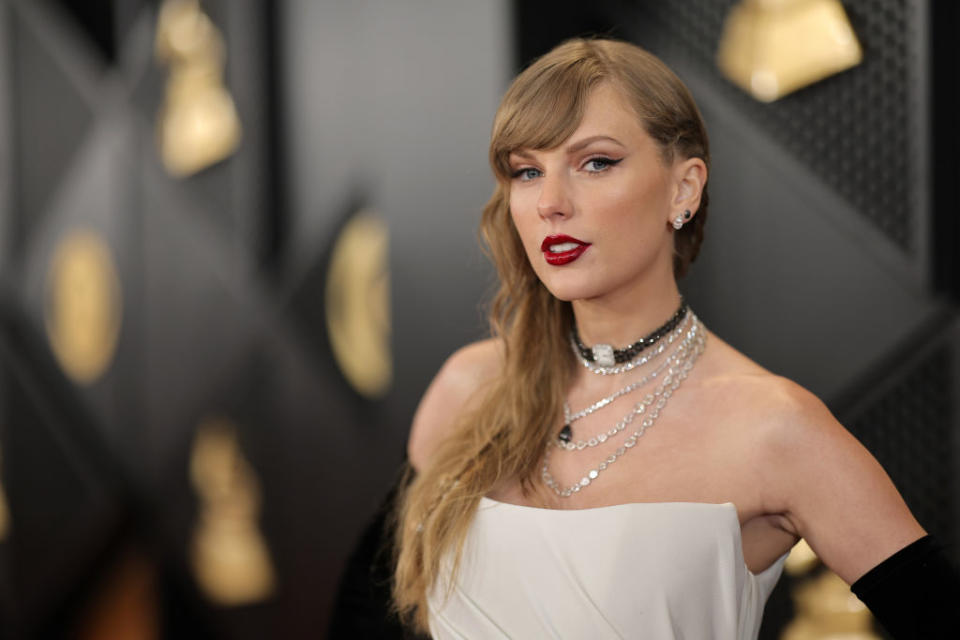 Taylor Swift poses on the red carpet in an elegant strapless dress, layered necklaces, and black gloves, with Grammy Award statues in the background