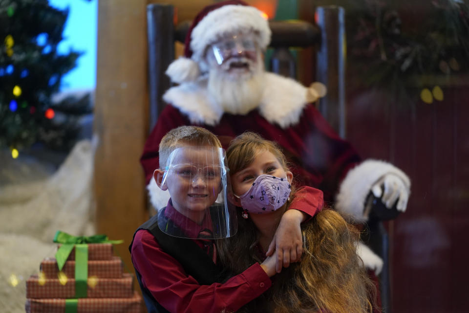 William Peargin, left, and Payton Peargin, right, both 8, pose for a socially distant photo with Santa Claus, who sits behind a sheet of plexiglass, at Bass Pro Shop in Rancho Cucamonga, Calif., on Dec. 4, 2020. In this socially distant holiday season, Santa Claus is still coming to towns (and shopping malls) across America but with a few 2020 rules in effect. (AP Photo/Ashley Landis)