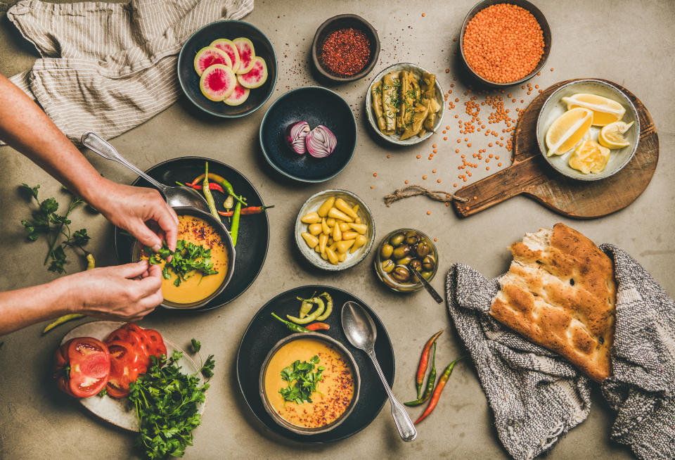 Flat-lay of turkish lentil soup Mercimek , vegetables, flatbread, pickled peppers and mans hands adding parsley to soup over concrete background.