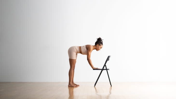 A Black woman wearing light colored shorts practices Half-Standing-Forward-Bend with a chair for support