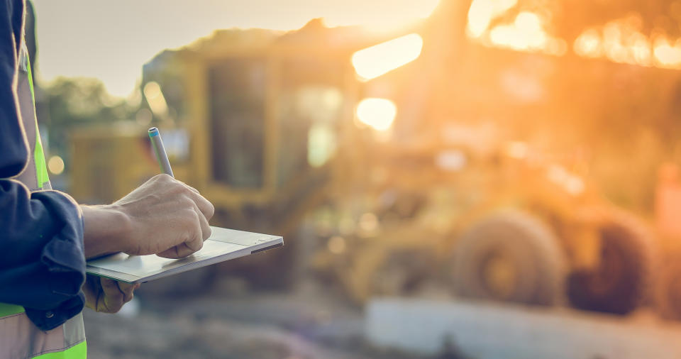 A person holding a tablet near a construction site with a bright sun in the background.