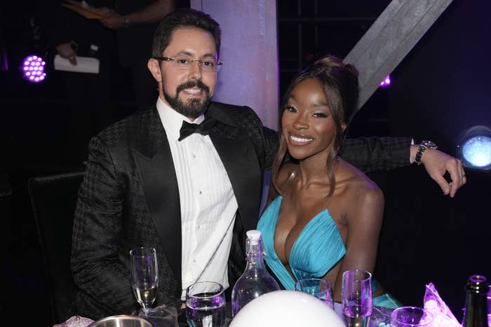 Jeff Lazkani in a tuxedo and a woman in a low-cut dress sit at a table with drinks and smile at the camera