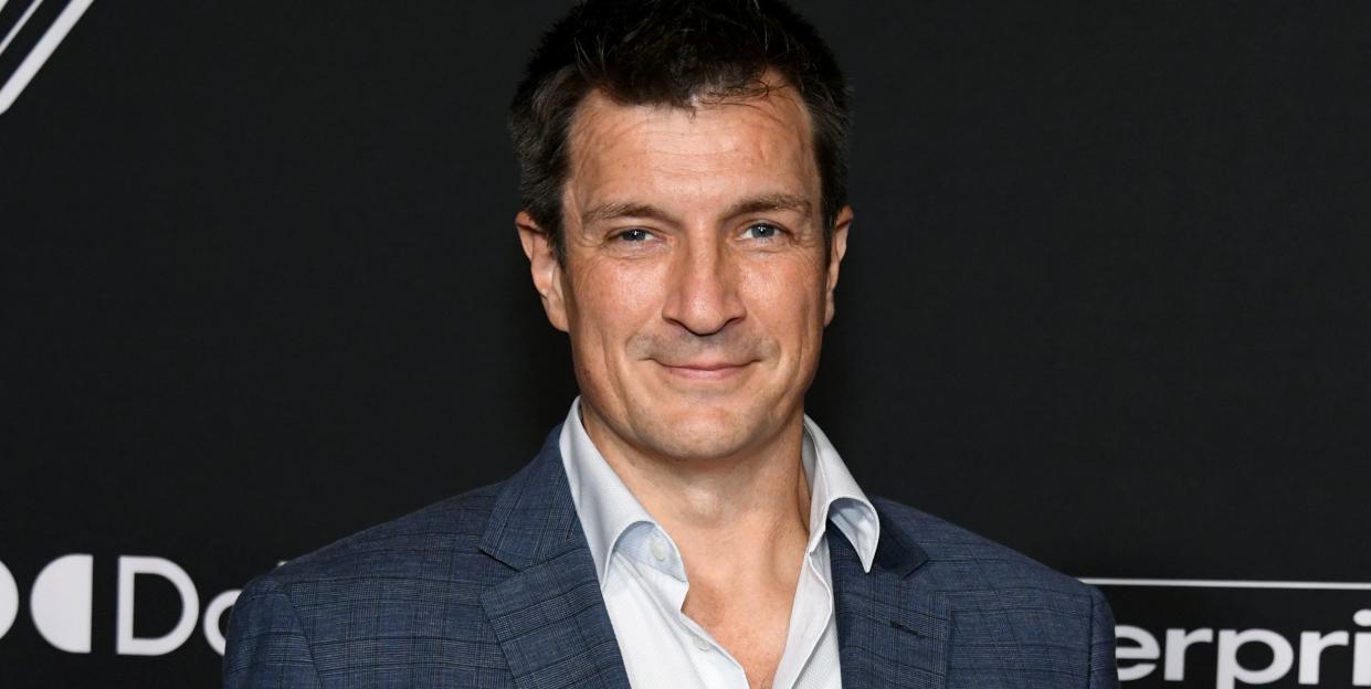 nathan fillion wearing a white collared shirt and a navy blue suit, standing in front of a black background at the world premiere of guardians of the galaxy volume 3 in april 2023