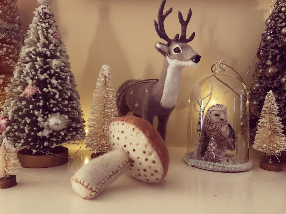 Close up of tiny, sparkling Christmas trees decorated with shiny silver and pink ornaments and glitter arranged neatly on a fireplace mantel. Tiny snow globe ornament with woodland owl figurine inside. Felt toadstool with glittering jewels. Reindeer. Festive holiday and celebration.