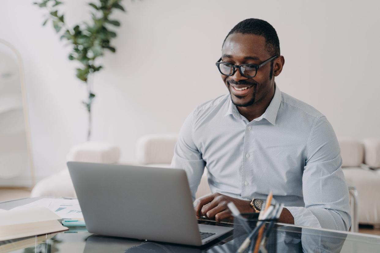 Smiling african american businessman wearing glasses working at laptop, makes successful deal. Happy black man employee glad to receive good news looking at computer screen at office desk.