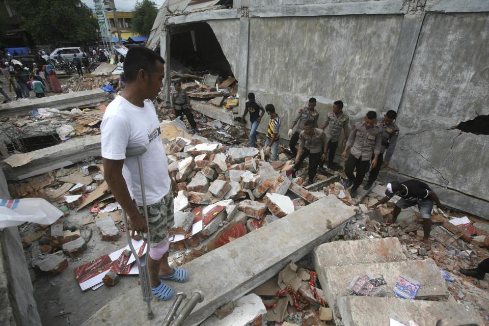 Faisal Marwan, left, who was injured in Wednesday's earthquake, stands on the ruins of his shop as police officers clear the rubble in Tringgading, Aceh province, Indonesia, Friday, Dec. 9, 2016. Over a hundred of people were killed in the quake that hit the northeast of the province on Sumatra before dawn Wednesday. Hundreds of people were injured and thousands buildings destroyed or heavily damaged. (AP Photo/Binsar Bakkara)