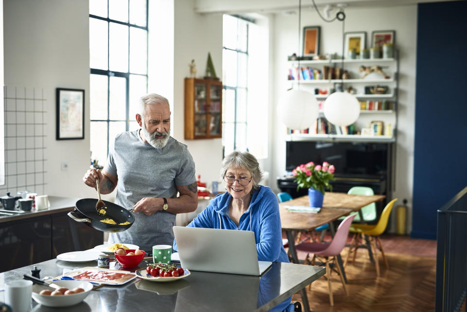 Retired couple in modern kitchen on the weekend, woman surfing the web, man holding frying pan and serving scrambled eggs