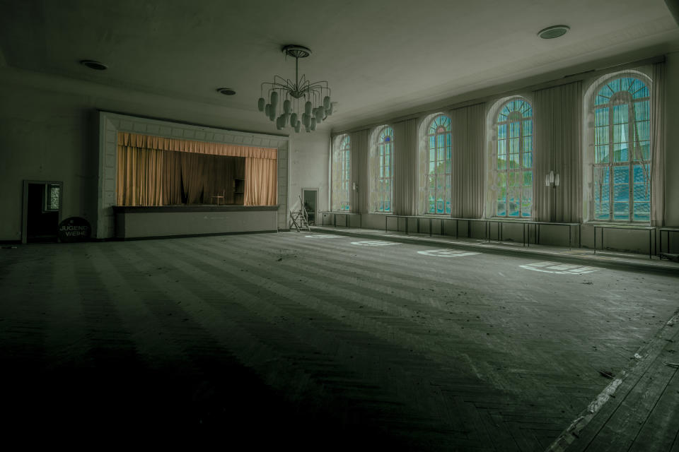 A large, empty, and dimly-lit auditorium with a stage and several tall windows. The chandeliers hang from the ceiling. No people are present