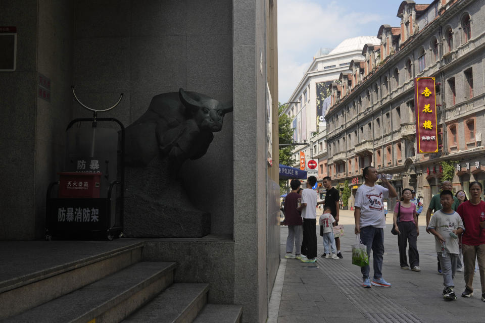 Visitors to a shopping district pass near a sculpture of a bull and anti-riot equipment outside a bank in Shanghai, Thursday, Aug. 1, 2024. (AP Photo/Ng Han Guan)