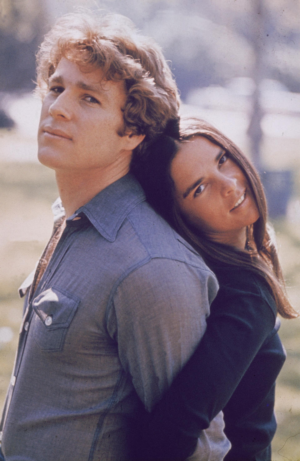 merican actors Ryan O'Neal and Ali MacGraw stand back to back outdoors in a still from the film, 'Love Story,' directed by Arthur Hiller, 1970.