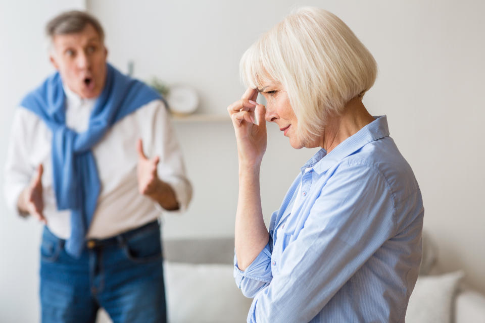 An older couple is having an argument. The woman in the foreground looks distressed with her hand on her forehead, while the man in the background gestures animatedly