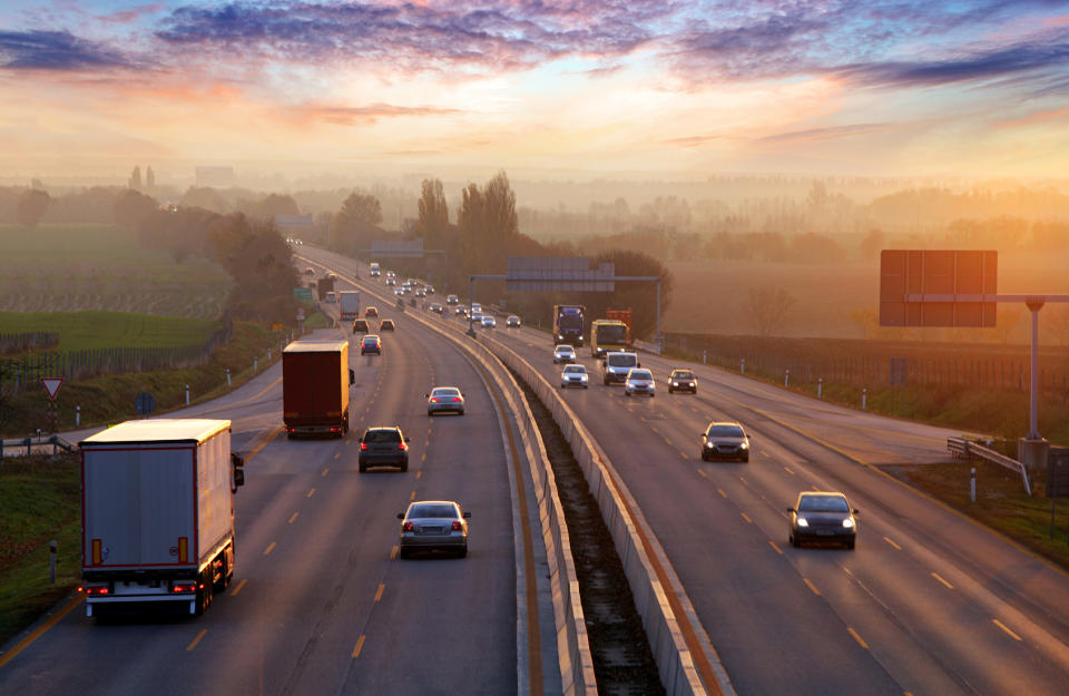 Highway traffic moves in both directions under a scenic sunset, with various vehicles including trucks and cars