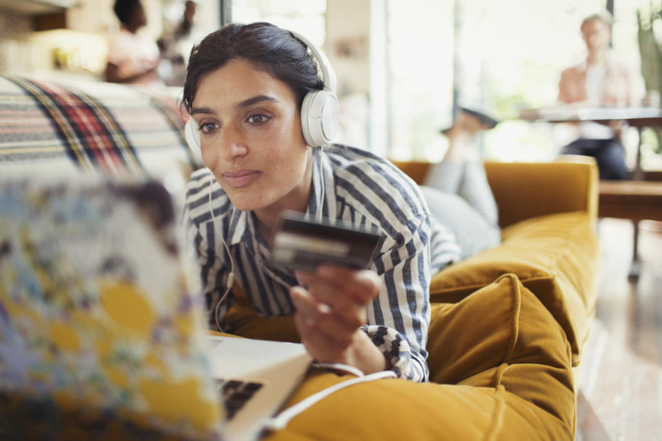 Young woman with credit card and headphones online shopping at laptop on living room sofa. Source: Getty