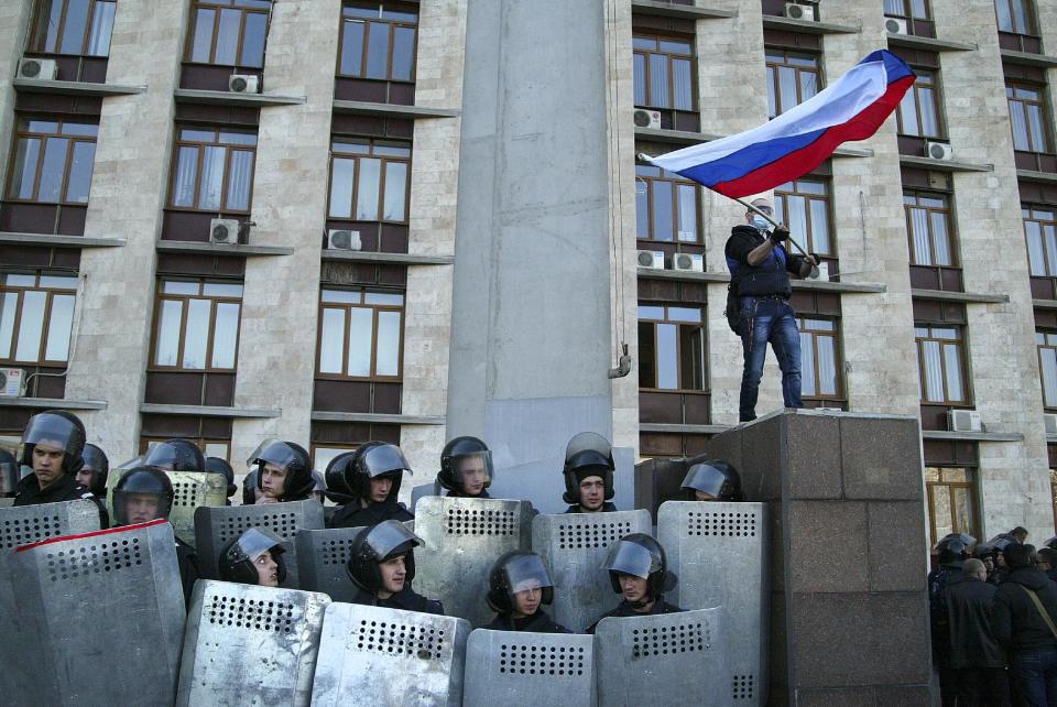Pro-Russian masked activist waves a Russian national flag above Ukrainian police at the regional administration building in Donetsk, Ukraine, Sunday, April 6, 2014. In Donetsk a large group of people surged into the provincial government building and smashed windows. A gathering of several hundred, many of them waving Russian flags, then listened to speeches delivered from a balcony emblazoned with a banner reading “Donetsk Republic.” (AP Photo/Alexander Ermochenko)
