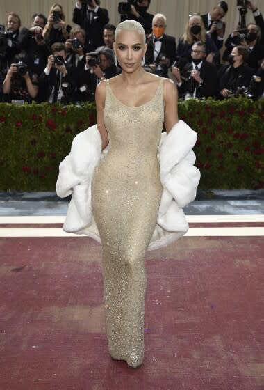 A woman stands on a red carpet posing in a sparkling gown and a white fur stole