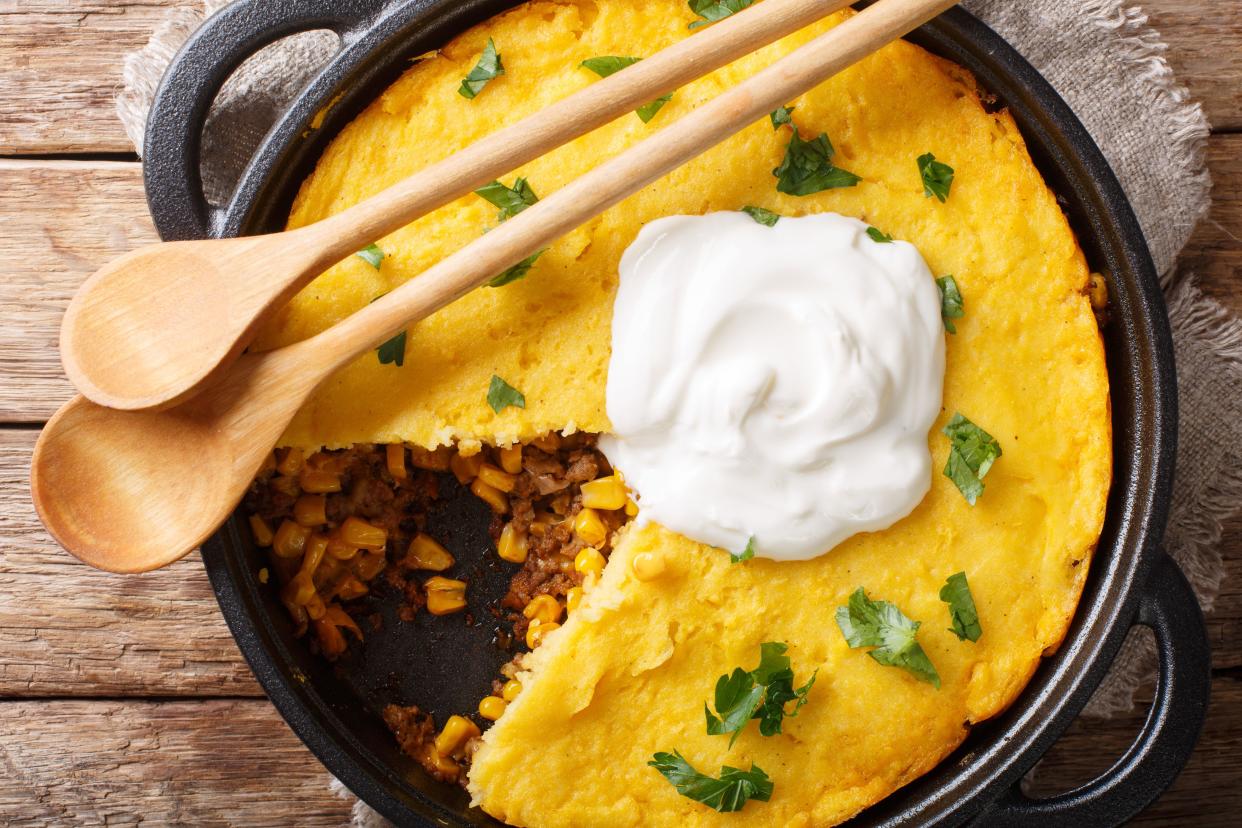 Top view of pork tamale pie in a round cast iron skillet, with two wooden spoons on top, one piece is missing, on a rustic natural wooden table