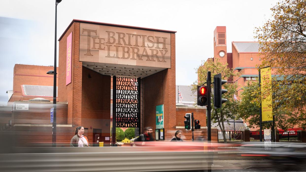  The exterior of The British Library, a sandstone building with "The British Library" carved above its entrance, with a metal gate moulded to say "British Library" repeatedly beneath it. Some people, blurred due to long exposure, walk in front of the library. 