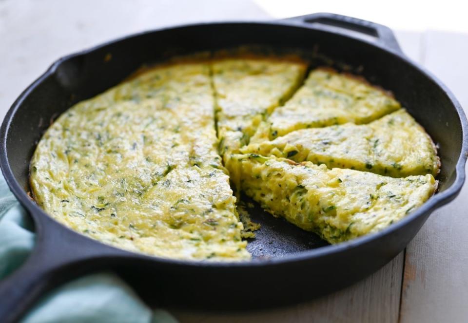 A cast iron skillet with a sliced, herb-infused frittata. The frittata has a fluffy texture and segments cut for serving