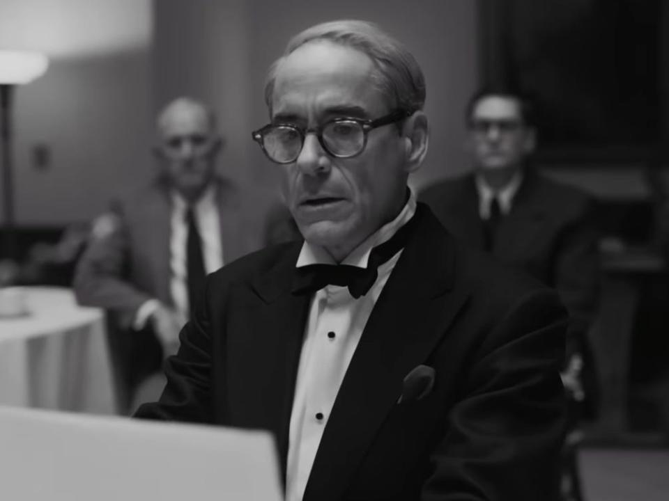 A black-and-white image of Robert Downey Jr. in "Oppenheimer." wearing a suit, bow tie, and glasses, with two male characters in the background.