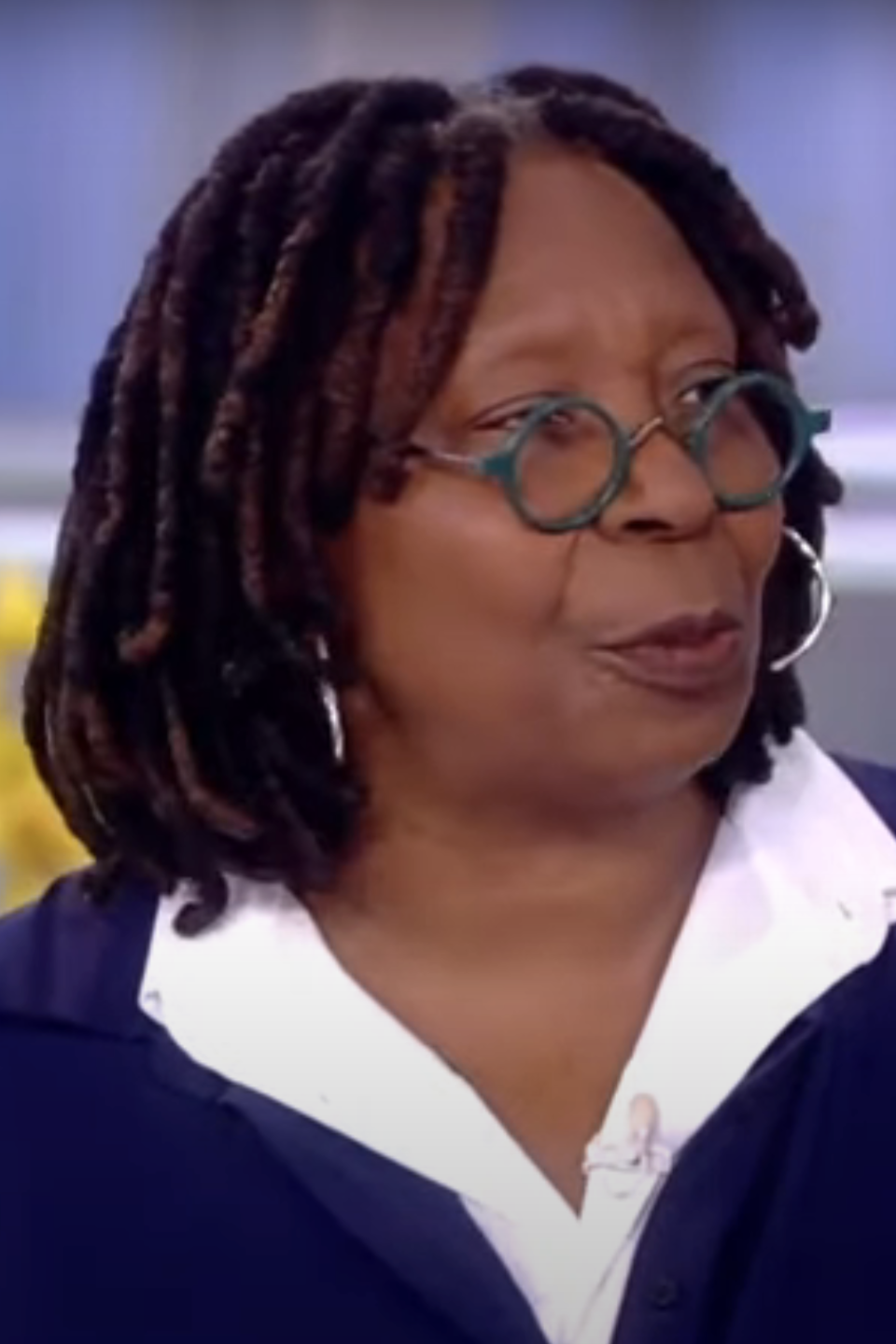 Whoopi Goldberg wearing glasses and a blue cardigan over a white top on a talk show