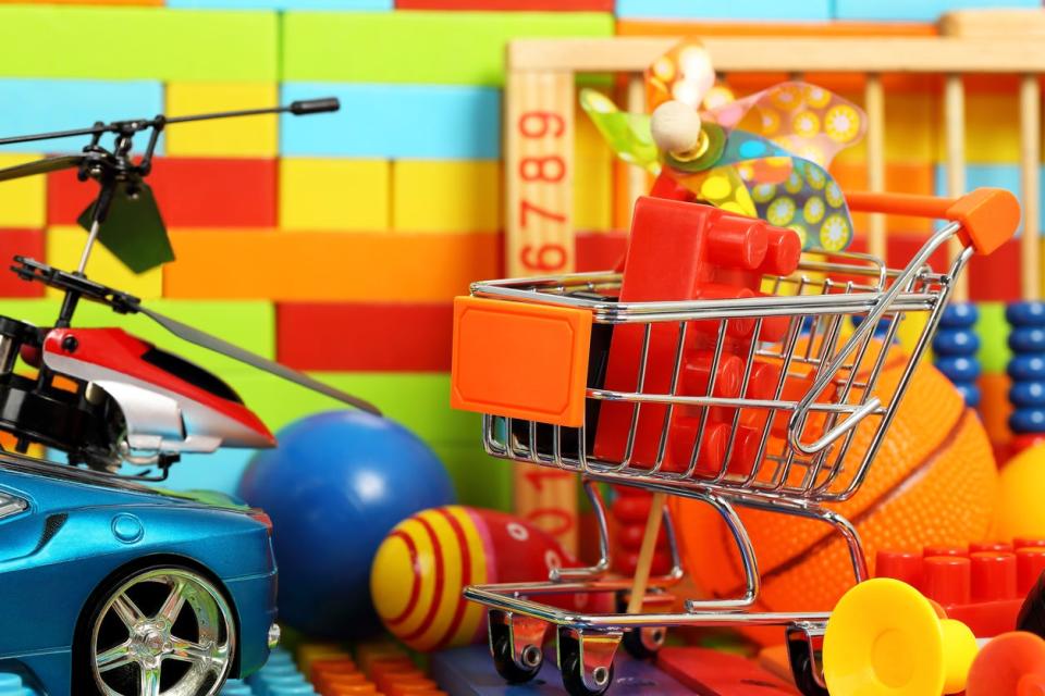 A mini shopping cart with a red lego inside a toy car and toy helicopter in front of a wall of colorful blocks