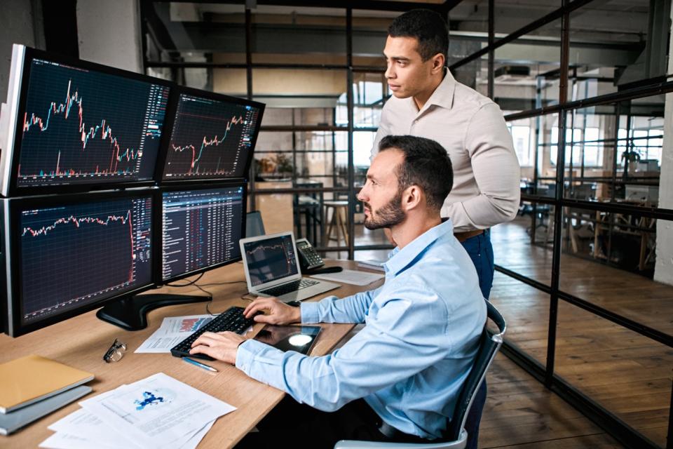 Two investors looking at four computer monitors with price charts on them.