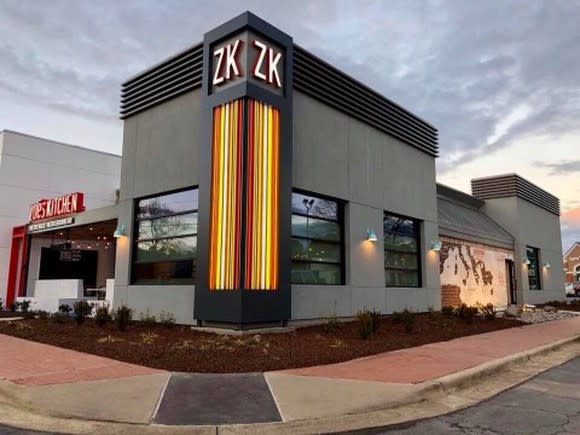 The outside of Zoe's Kitchen's new store in North Carolina. The building is modern minimalist, with orange lighting running up the corner of the building, and a new ZK logo at the top.