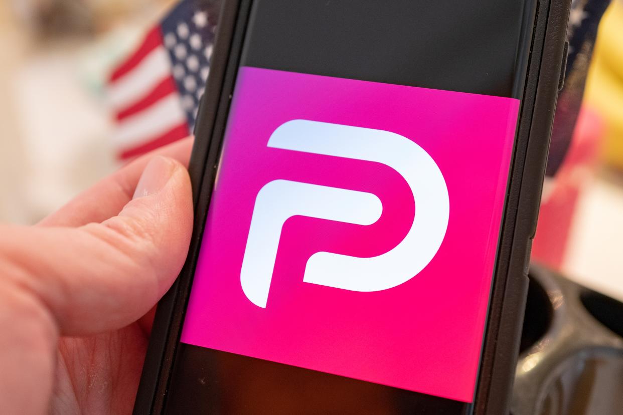 Illustrative image of human hand with mobile device showing logo for the social media platform Parler with American flag visible in background.