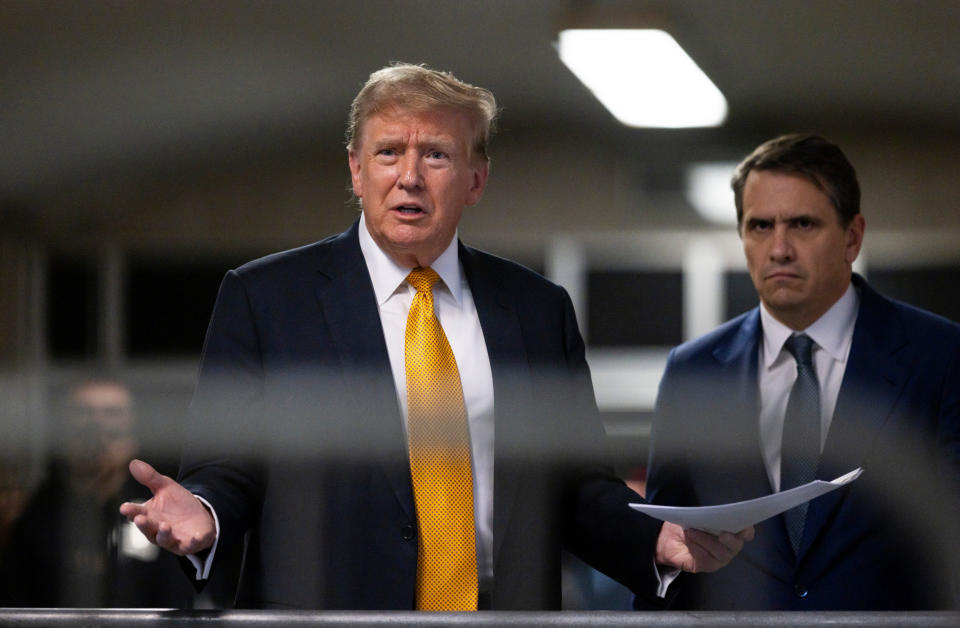 Donald Trump speaks while holding documents, accompanied by another man in a suit with a serious expression