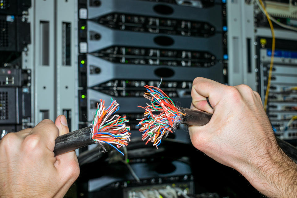 Two hands holding up two roughly cut network cables in front of a rack of enterprise routers.