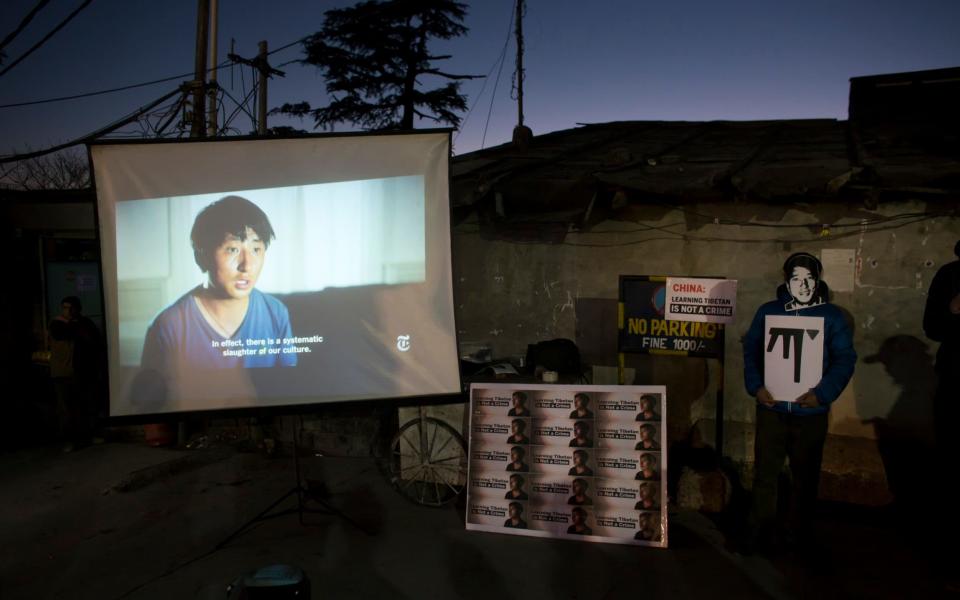 An exile Tibetan wearing a mask in the likeness of 32-year-old Tashi Wangchuk, stands next to a screen projecting a New York Times video during a street protest demanding his release - AP