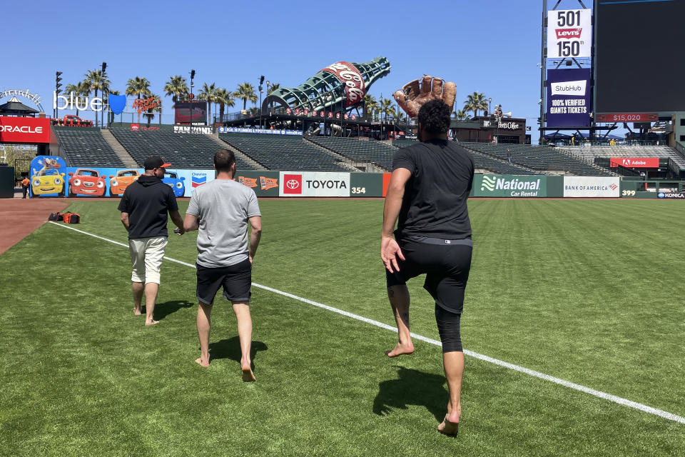 FILE - From left to right, San Francisco Giants assistant pitching coach J.P. Martinez, human performance coach Harvey Martin and pitcher Sean Manaea walk barefoot toward the center field bullpen for a breathing session before a baseball game against the Baltimore Orioles in San Francisco, June 3, 2023. Giants star Joc Pederson is scared to fly and shortly after arriving in San Francisco he began working with the club's director of mental health and wellness Shana Alexander and human performance specialist Martin to cope with his flight anxiety. (AP Photo/Janie McCauley, File)