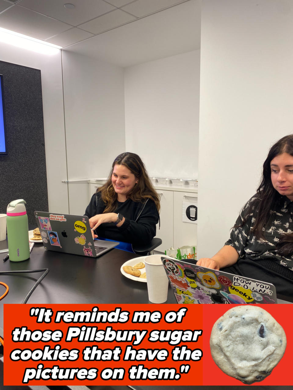 Two people are sitting at a table in a conference room with laptops covered in stickers beside plates of food and cookies