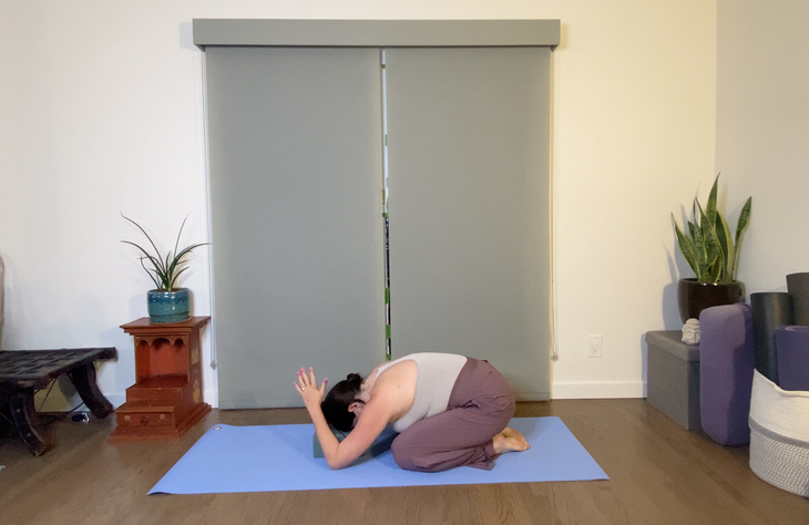 A woman folds over into Child's Pose. She is wearing a gray tank and purple pants and practices on a blue mat, There is a curtain behind her.