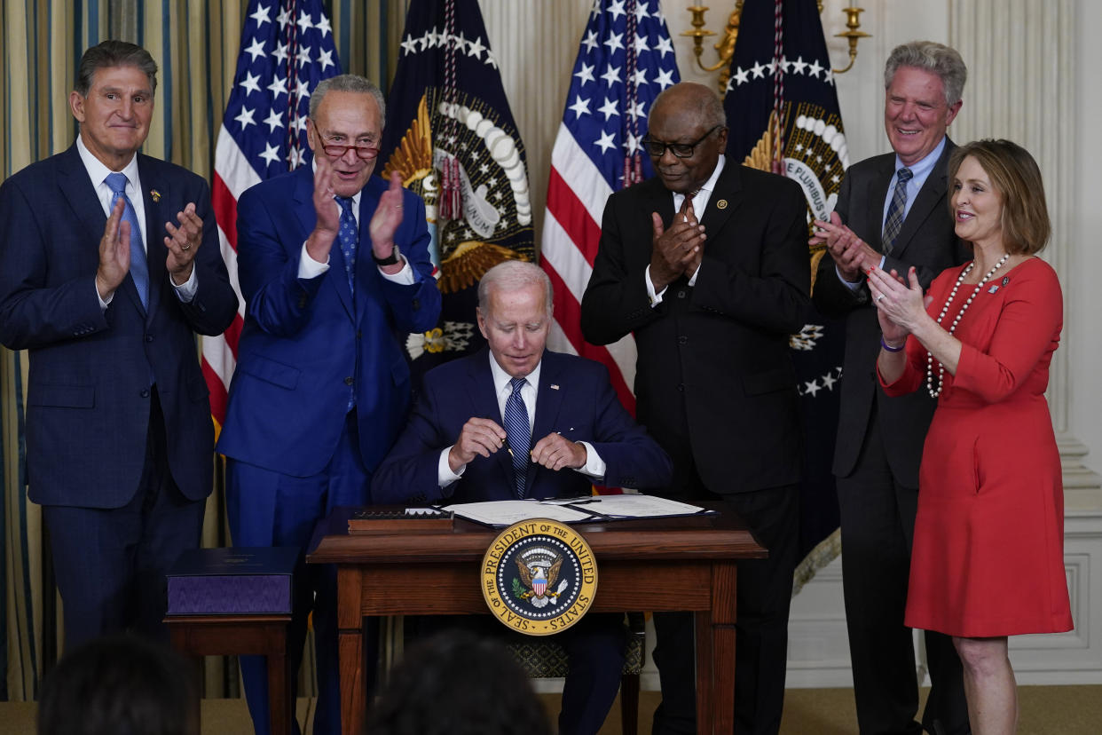 President Biden sits at a small desk bearing a presidential seal while Sen. Joe Manchin, Senate Majority Leader Chuck Schumer, Rep. James Clyburn, Rep. Frank Pallone and Rep. Kathy Castor stand behind him, appearing to applaud. 