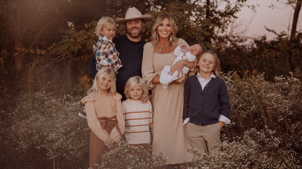 Family of five in neutral colors standing close together and smiling with greenery in the background