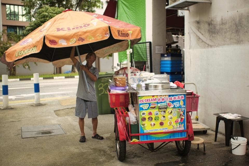An elderly Singaporean man with a slight frame holds a giant umbrella next to his ice cream cart.