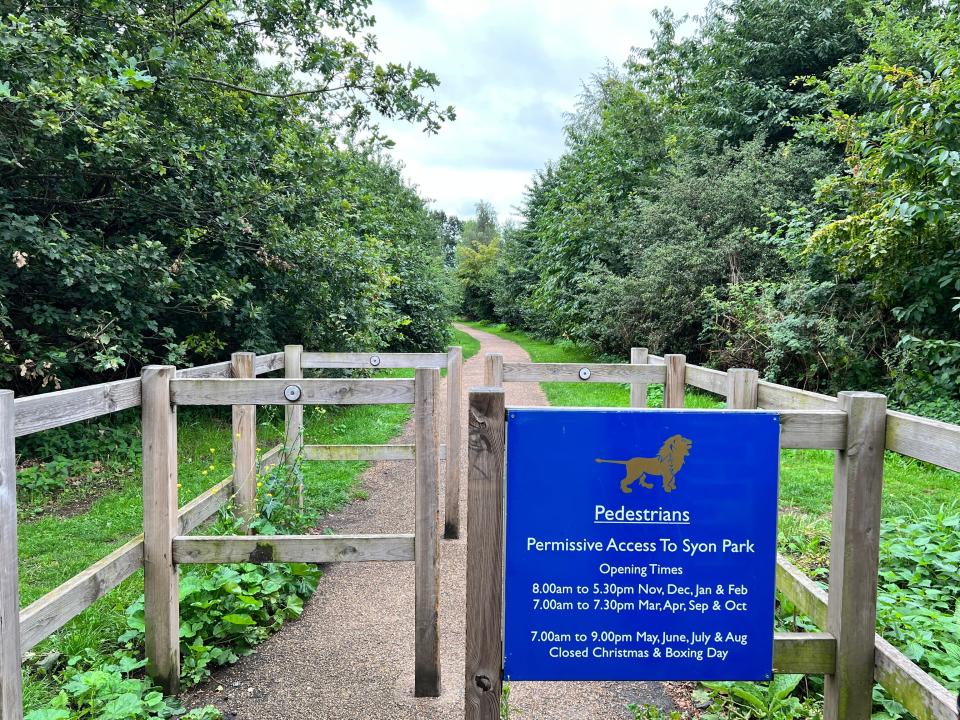 A blue sign displays the opening times for Syon Park, attached to a short wood fence with a path extending through greenery and trees in the background