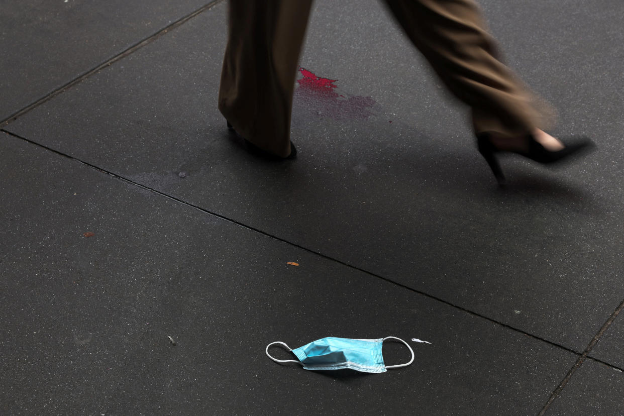 A blue surgical face mask lies on the sidewalk as the legs of a woman are seen walking by.